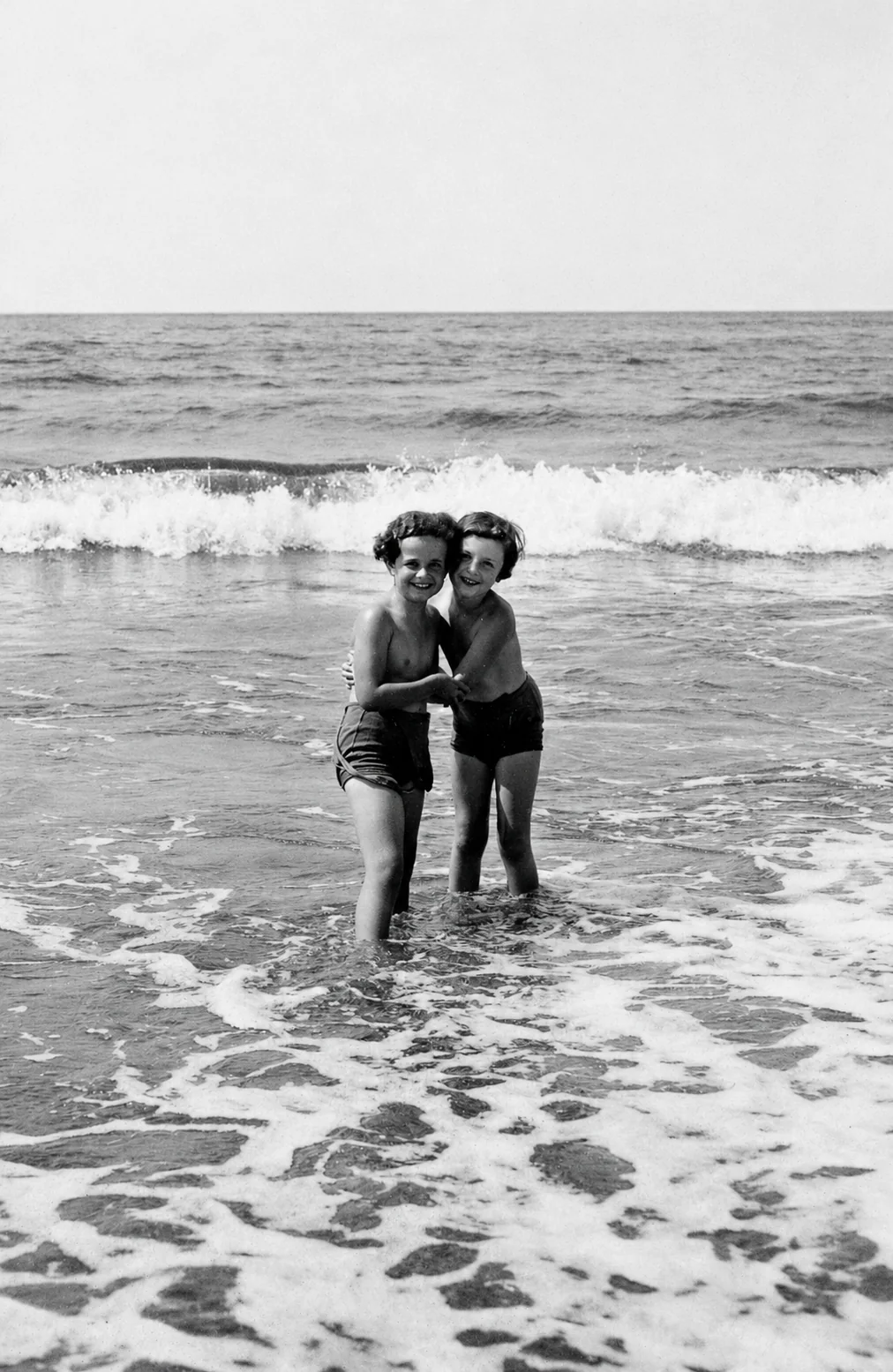 Dagmar Hirschberg and Gaby Kopelman, two young girls in swimming costumes standing arm in arm in the surf, laughing, with a wave breaking behind them. 1933.