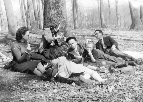 A family picnic among bare trees in the 1930s: two women, two men and two girls sitting on the leaves, eating.